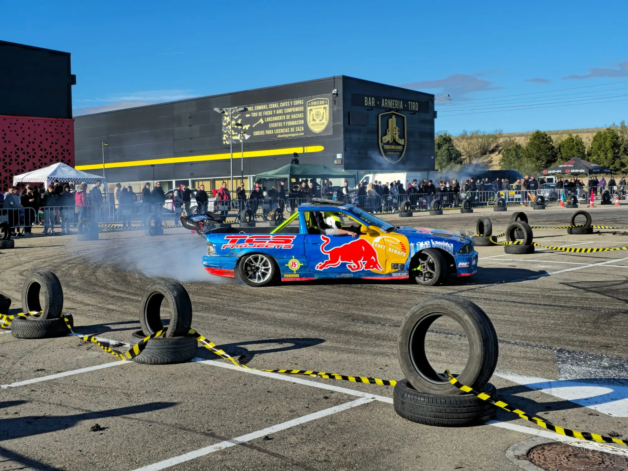 BMW de drift con decoración de Red Bull quemando rueda entre neumáticos, con el público detrás de las vallas